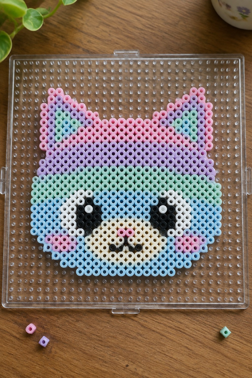 Colorful perler beads forming a cat face on clear pegboard with loose beads.