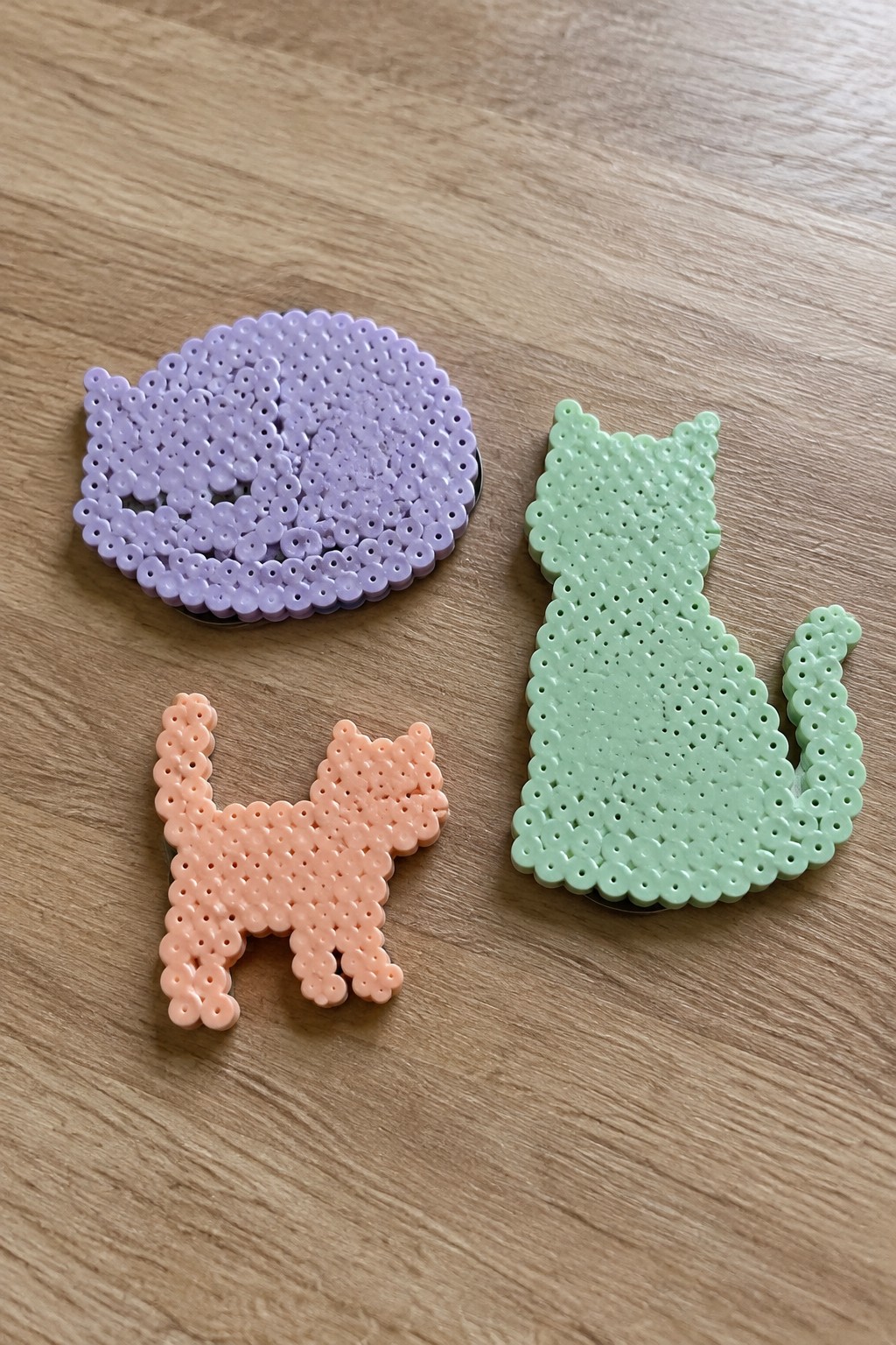 Three Perler bead cat designs in purple round, peach standing, and green standing silhouettes on wood.