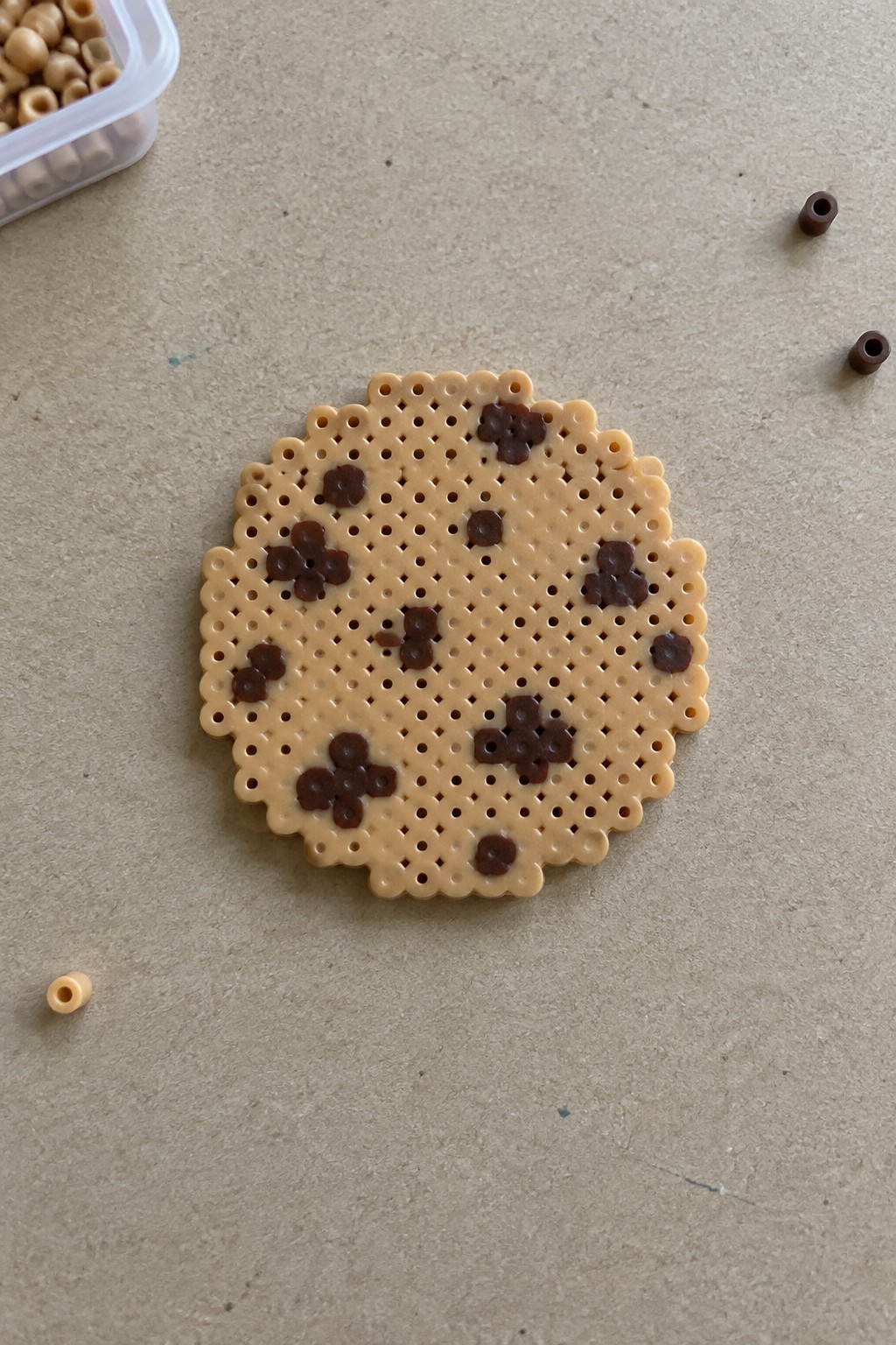 Perler bead cookie pattern with tan base and scattered brown chocolate chip clusters on a pegboard.