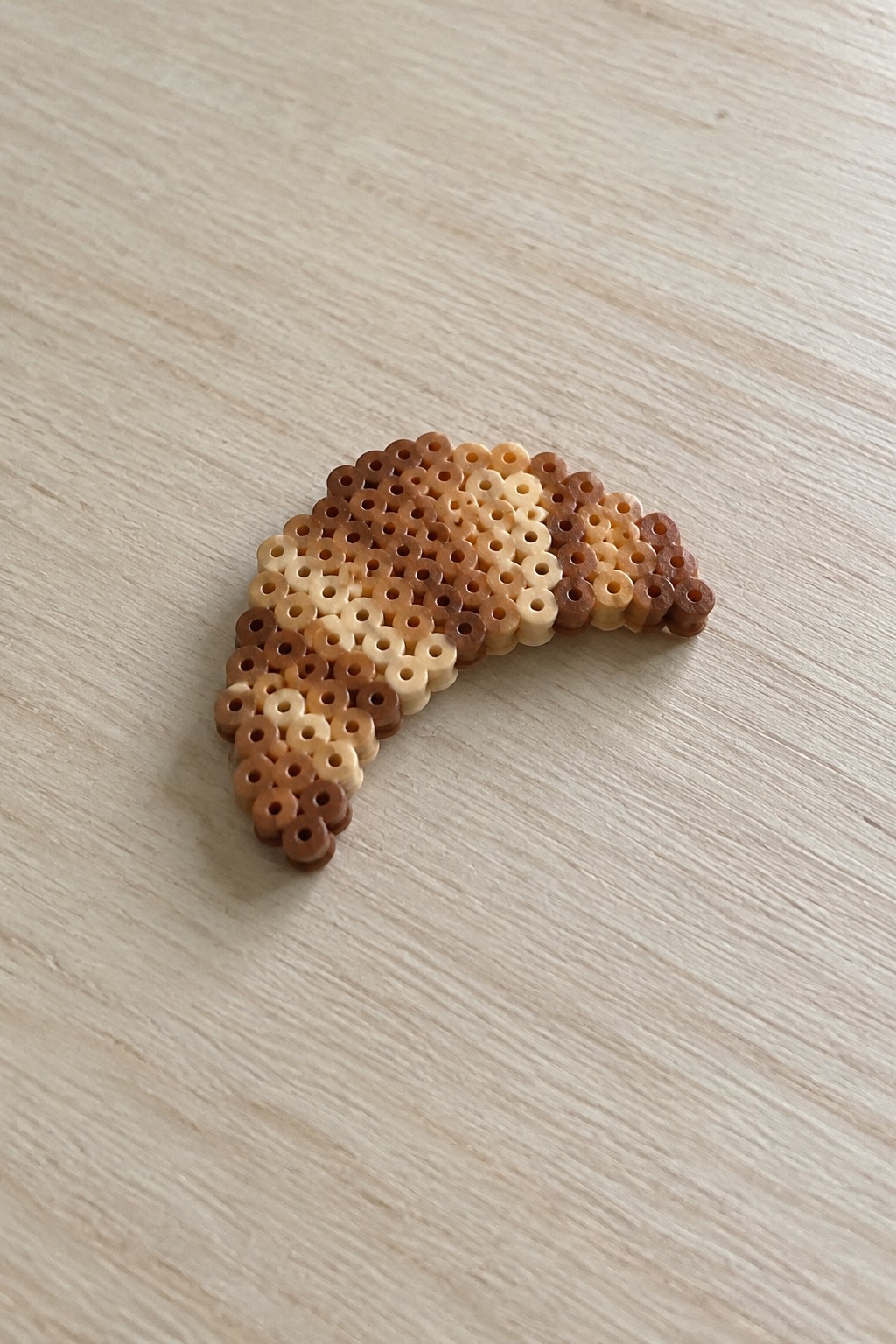 Beaded croissant craft with gradient brown perler beads on wooden table.