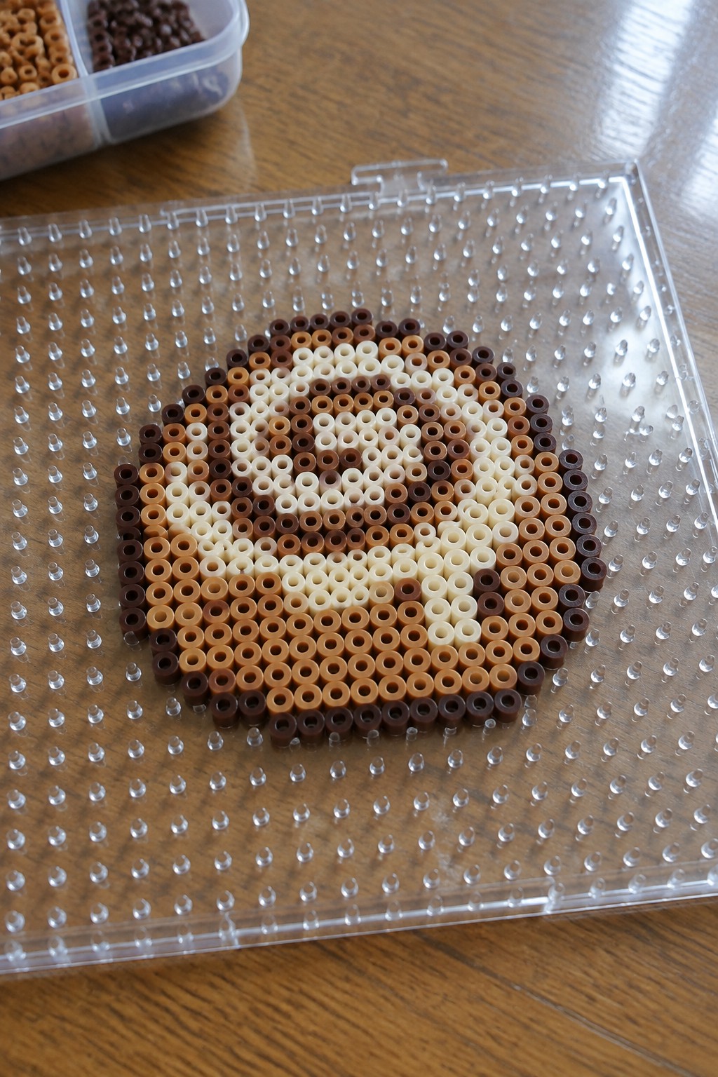 Brown perler beads forming cartoon animal face on clear pegboard with bead container nearby