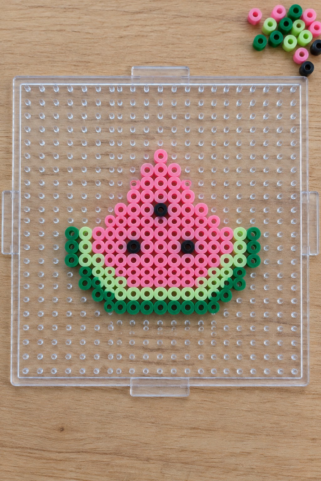 Pink and green perler bead watermelon with black seeds on clear pegboard.