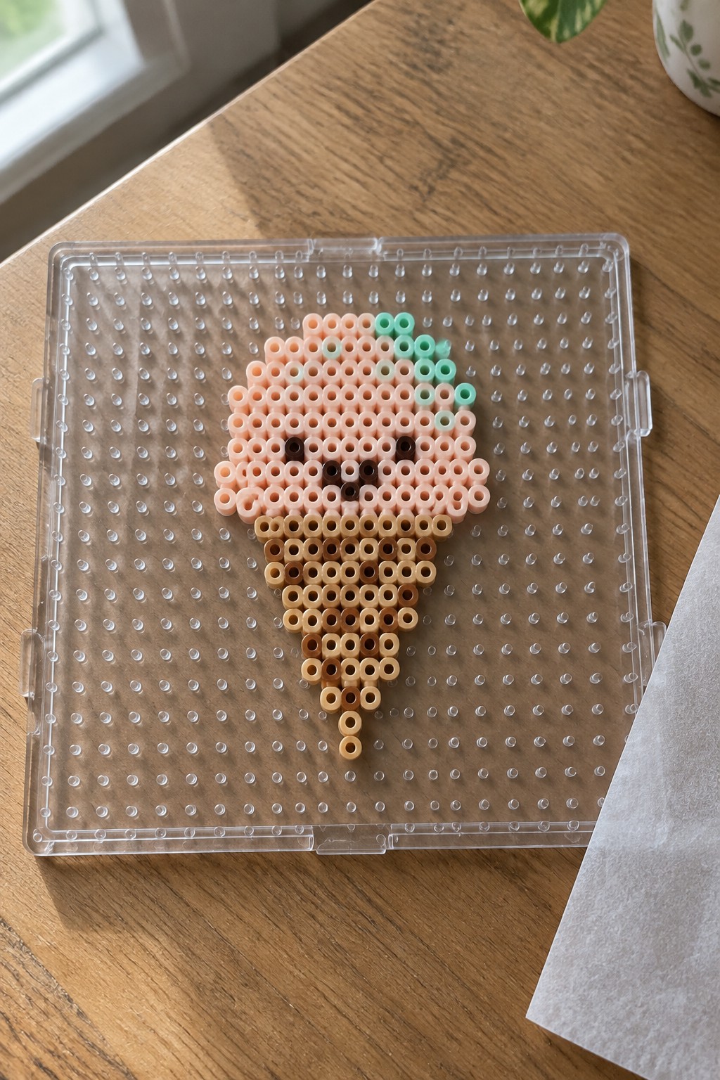 Perler bead ice cream cone on a pegboard with a pink scoop, mint green accents, brown facial details, and a brown cone.