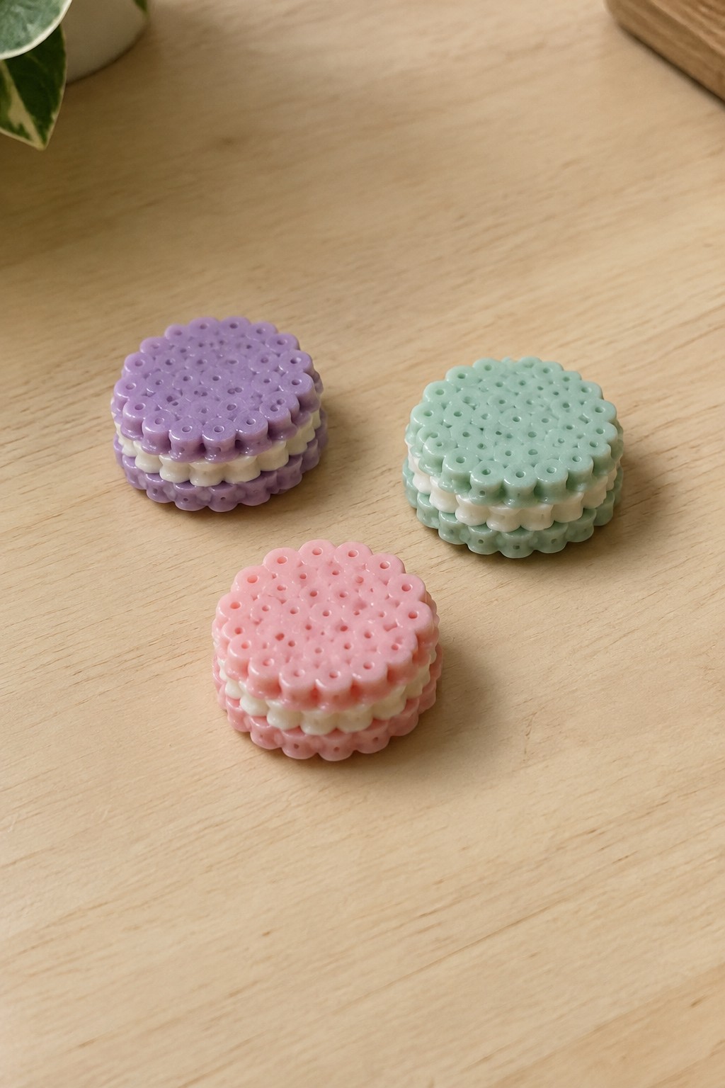 Three pastel perler bead macarons in purple, green, and pink on wood.