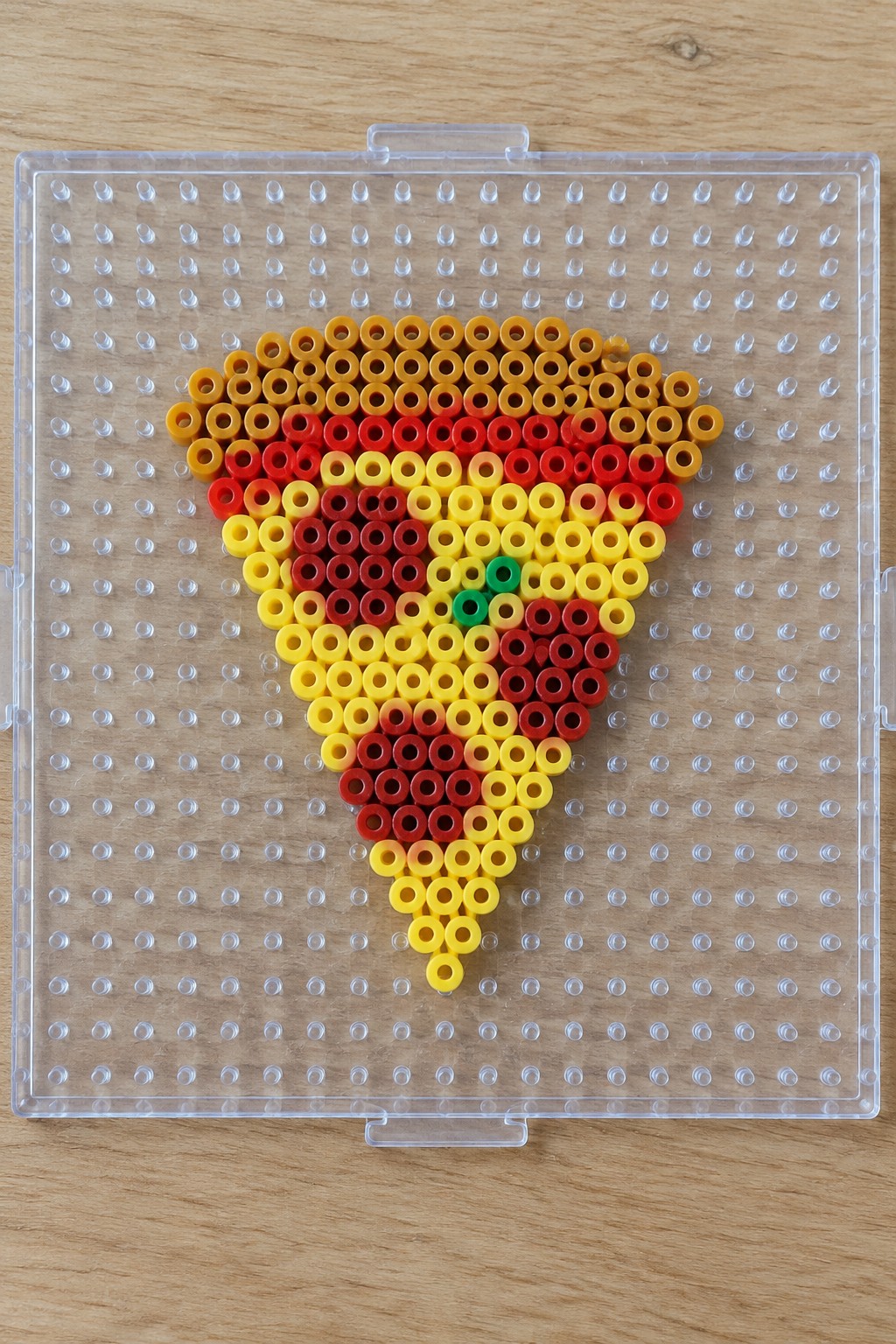Perler beads forming a colorful pizza slice on a clear pegboard over wood.
