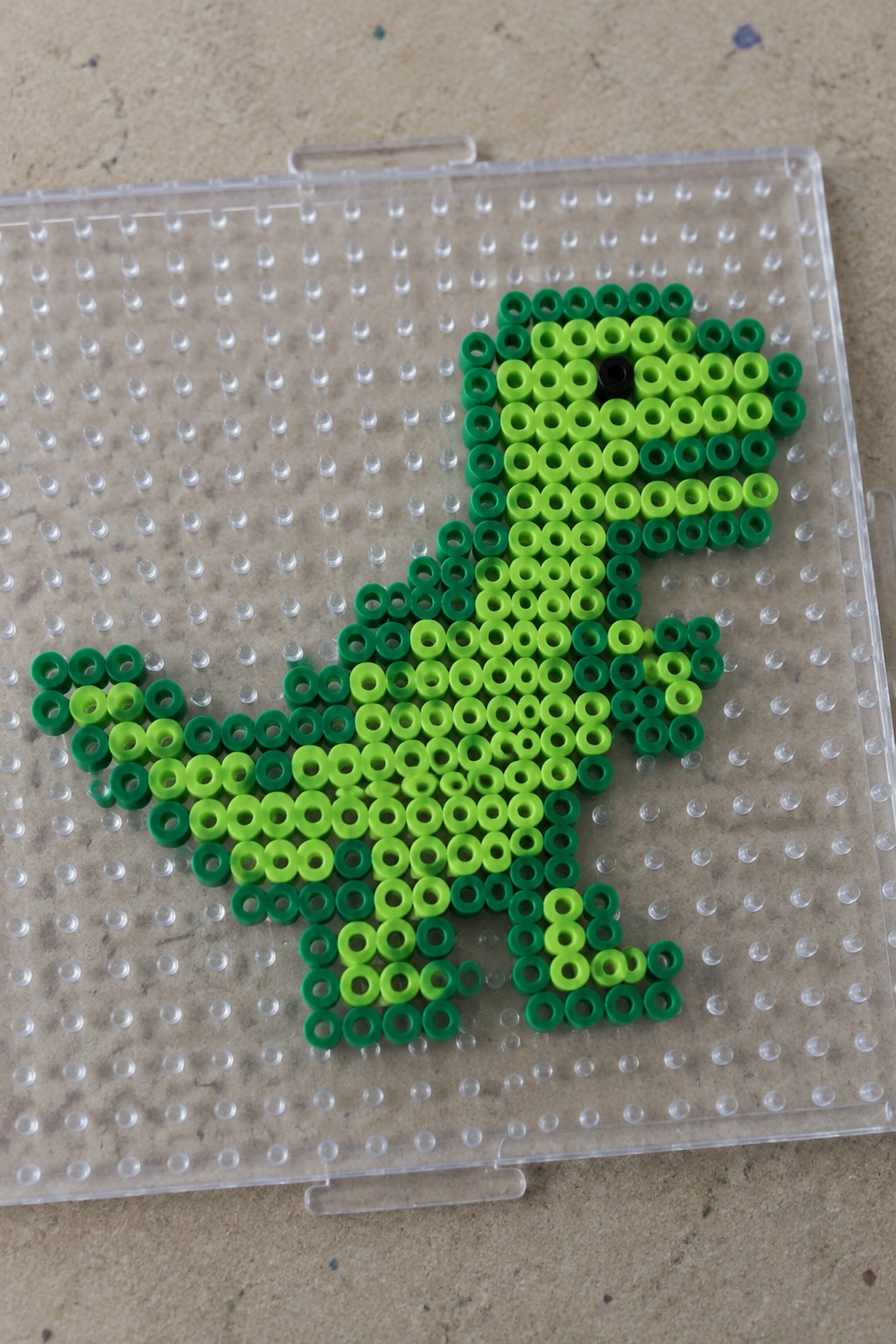 Green Perler beads form a T-Rex dinosaur on clear pegboard.