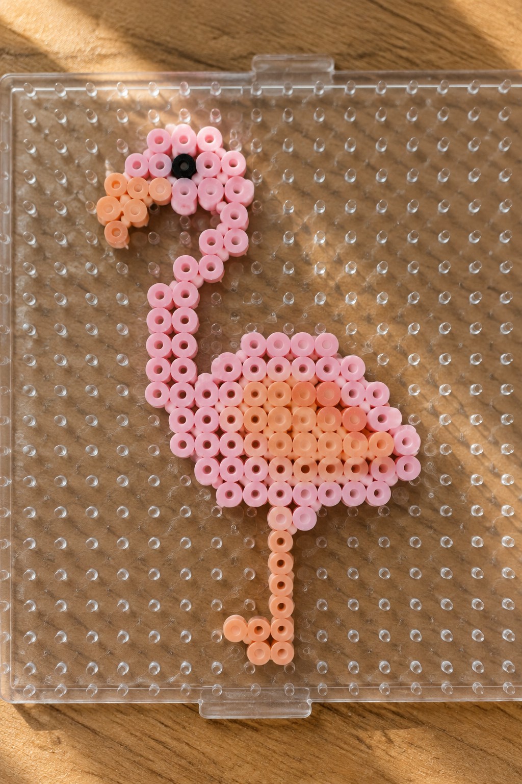 Pink perler bead flamingo with black eye on clear pegboard over wood