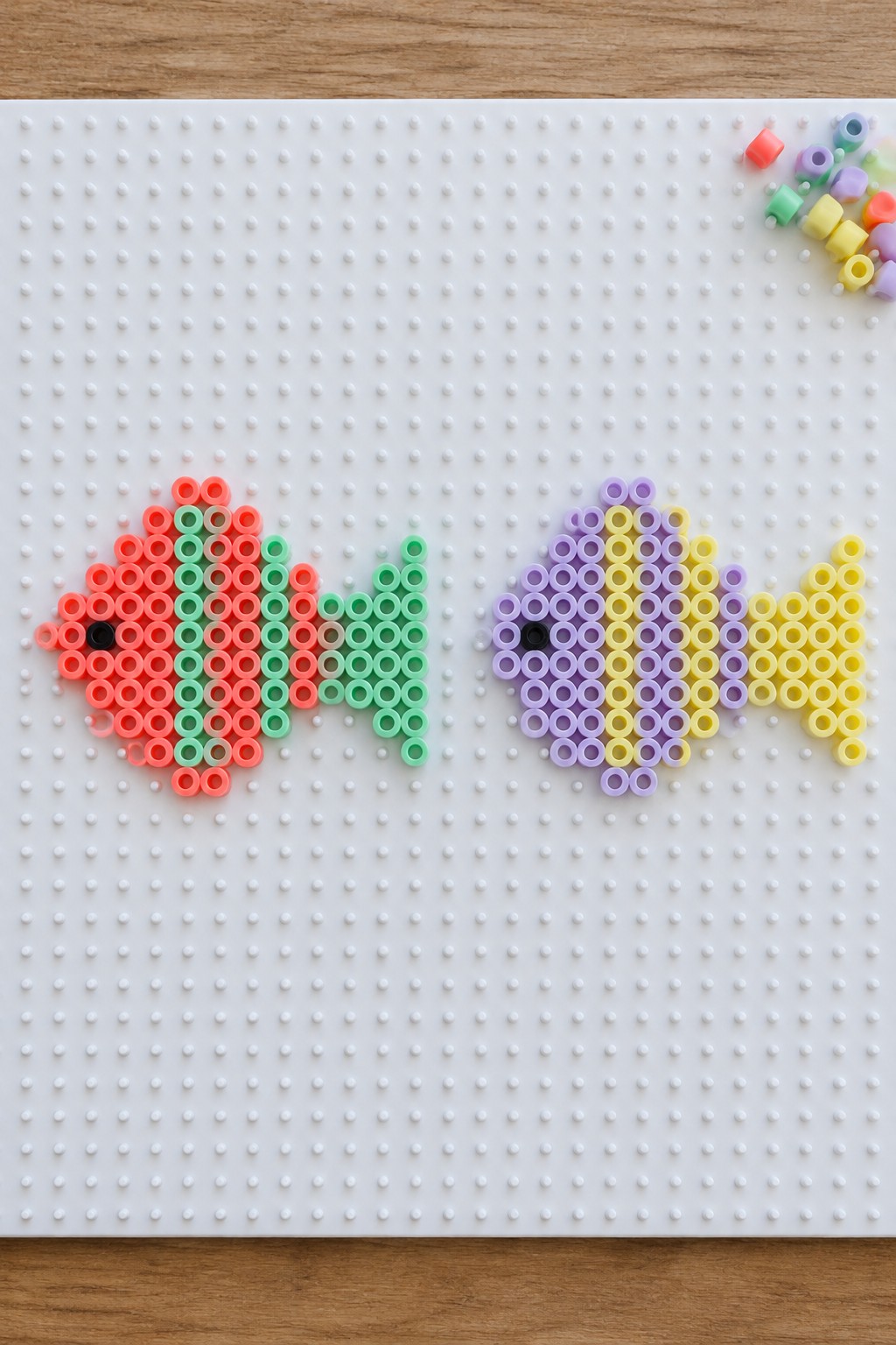 Two perler bead fish on white pegboard, red-green and purple-yellow with loose beads nearby