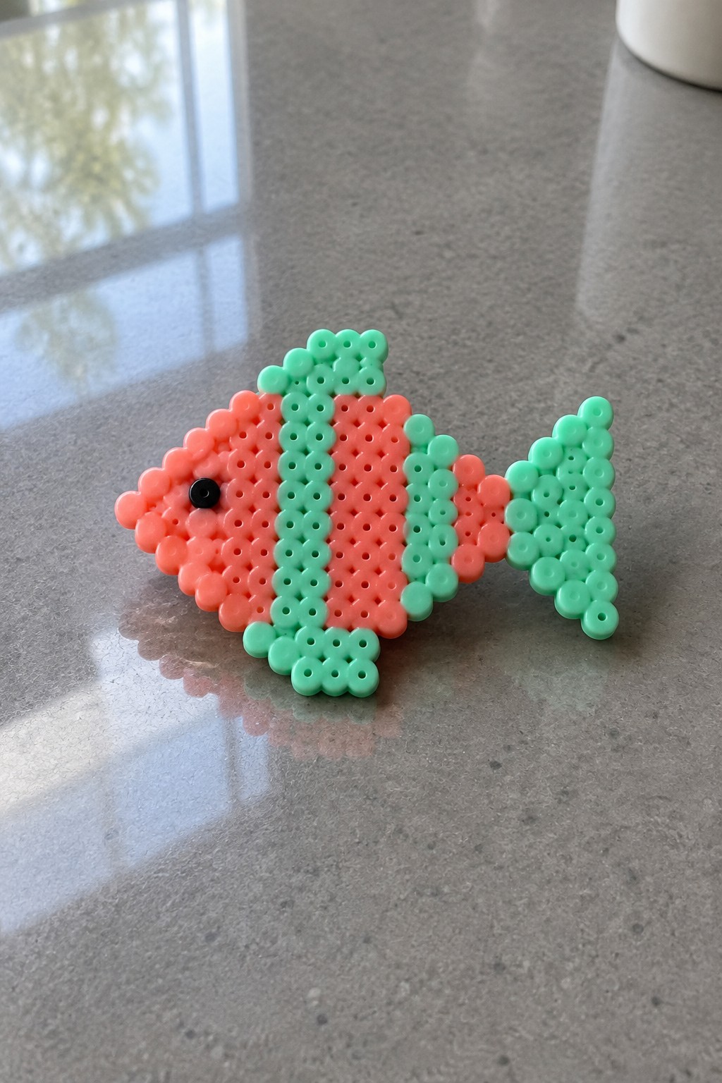 Perler bead fish pattern on a pegboard using coral and mint green beads with a black eye bead.