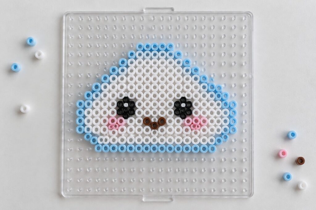 20 Kawaii Perler Bead Patterns With Expressive Faces Perler beads forming a cartoon face with blue border, black eyes, and pink cheeks on pegboard.