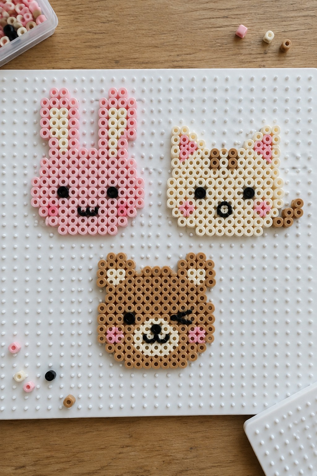 Three unfused Perler bead animal faces on a pegboard: a pink bunny, a cream cat, and a brown bear with a wink.