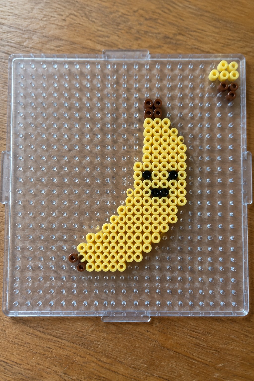 Yellow perler bead banana with black face and brown stem on pegboard