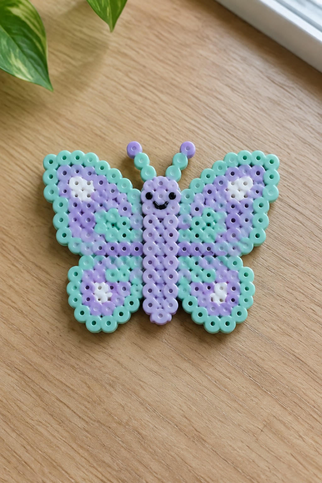 A Perler bead butterfly on a pegboard with mint green borders, purple and lavender wing sections, white accents, and a kawaii face on the body.