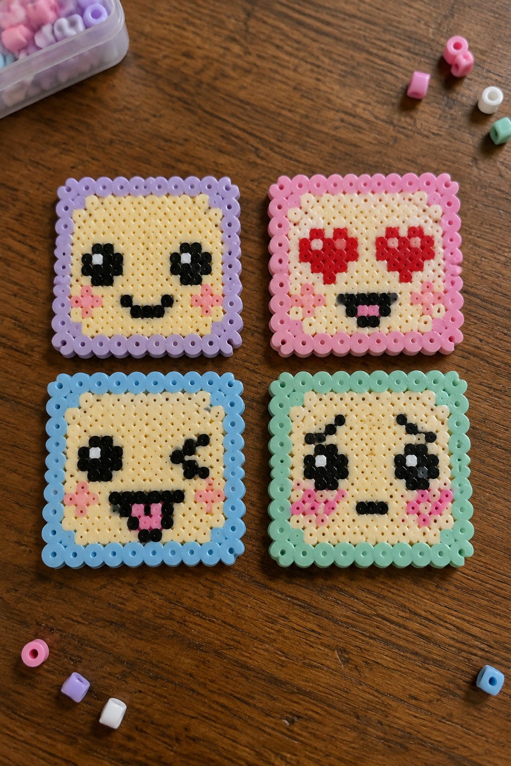 Four perler bead emoji faces with purple pink blue green borders on wood table
