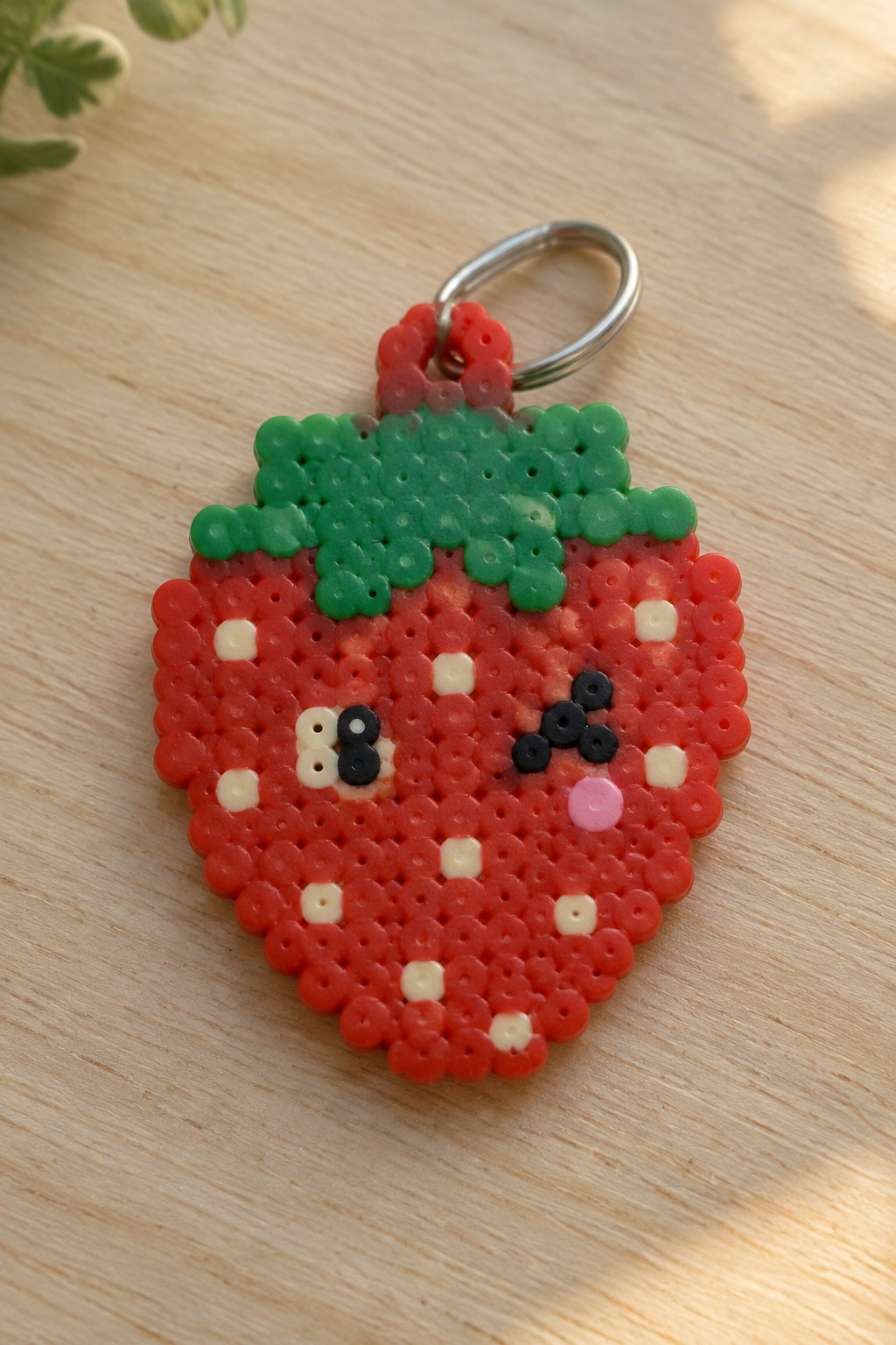 Fused Perler bead strawberry keychain with red body, green leaves, black and white eyes, and pink cheek attached to a metal ring.