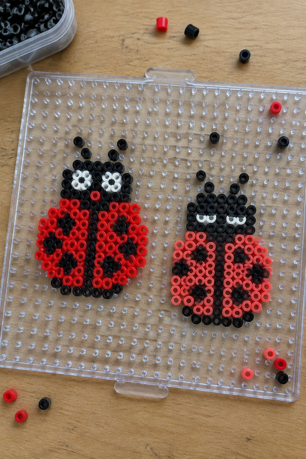 Two perler bead ladybugs in red and pink on a clear pegboard.