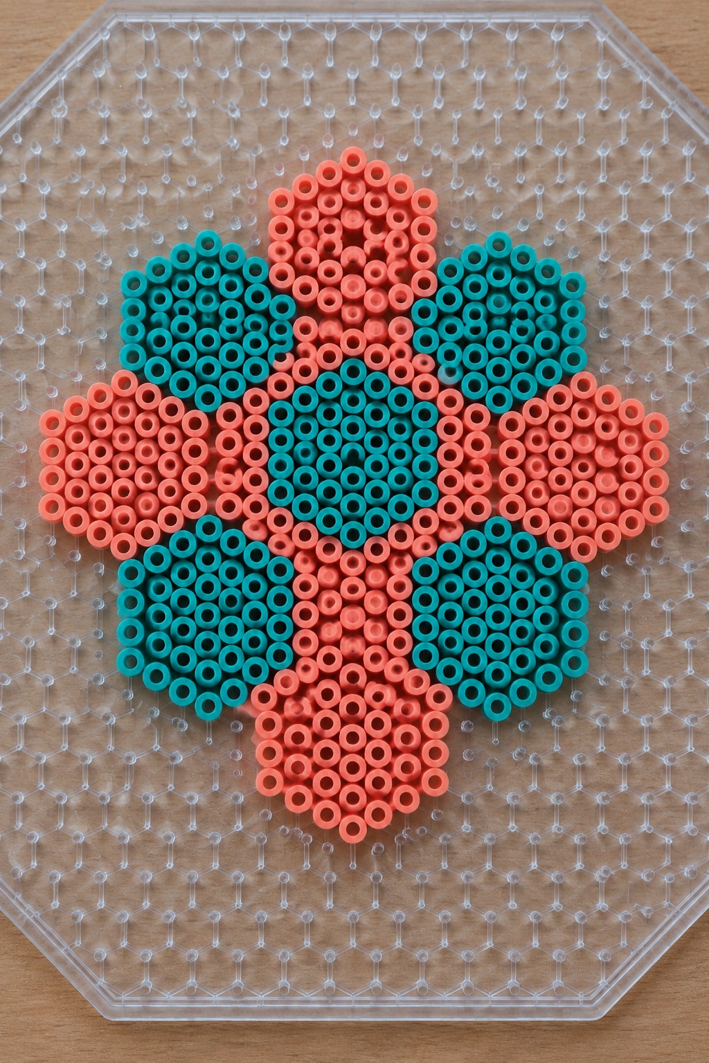 Coral and teal perler beads form a flower pattern on a clear pegboard.