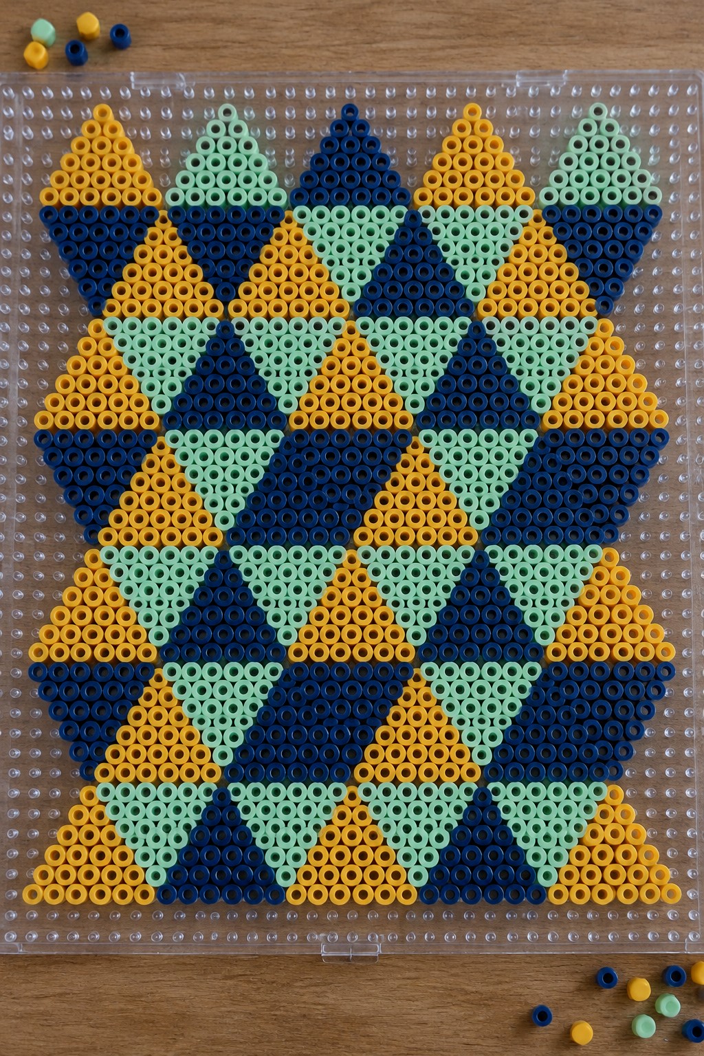 Perler beads on a pegboard forming a large symmetrical geometric pattern in yellow, navy blue, and mint green.