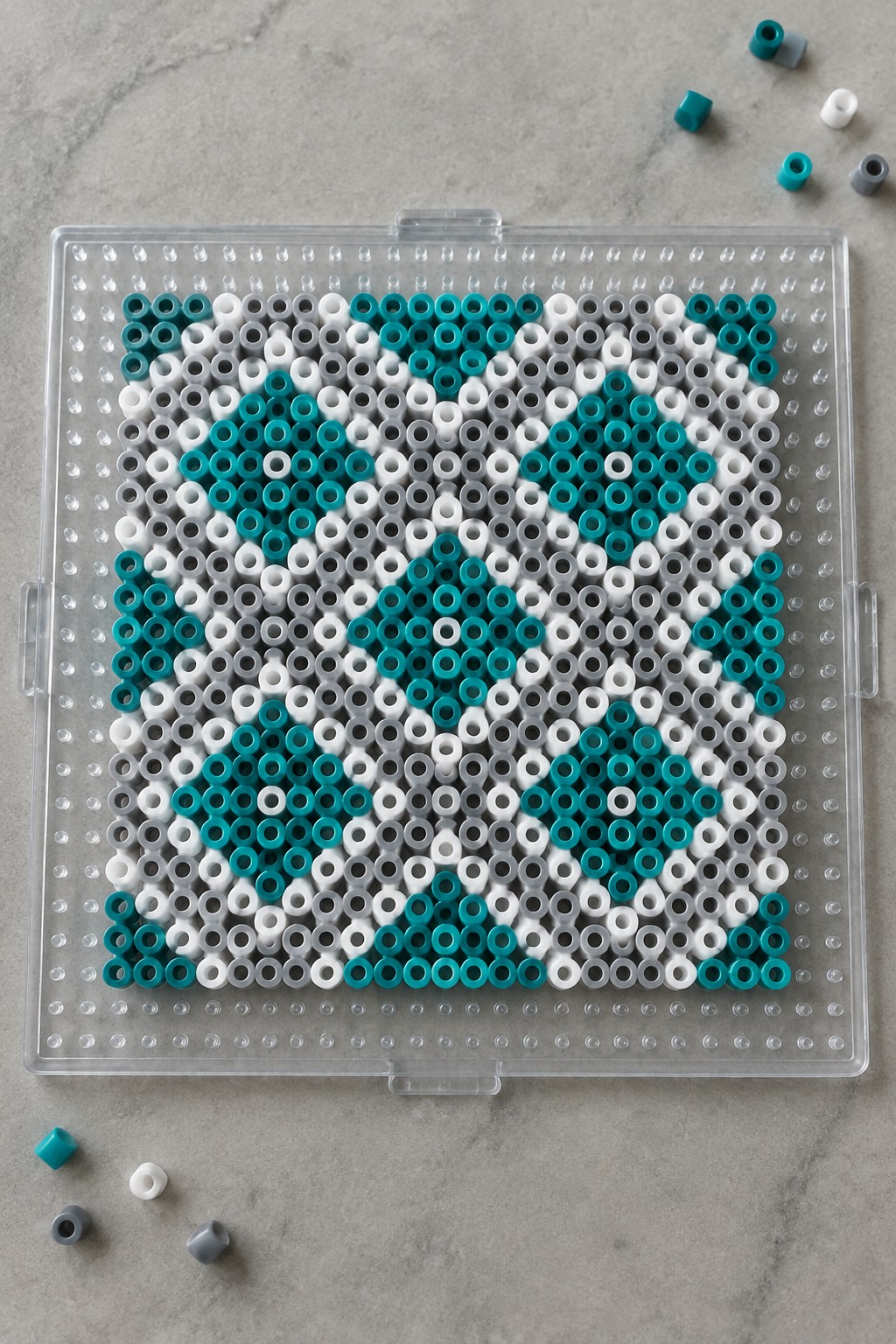 Unfused Perler beads forming a teal, white, and gray geometric diamond pattern on a clear pegboard.
