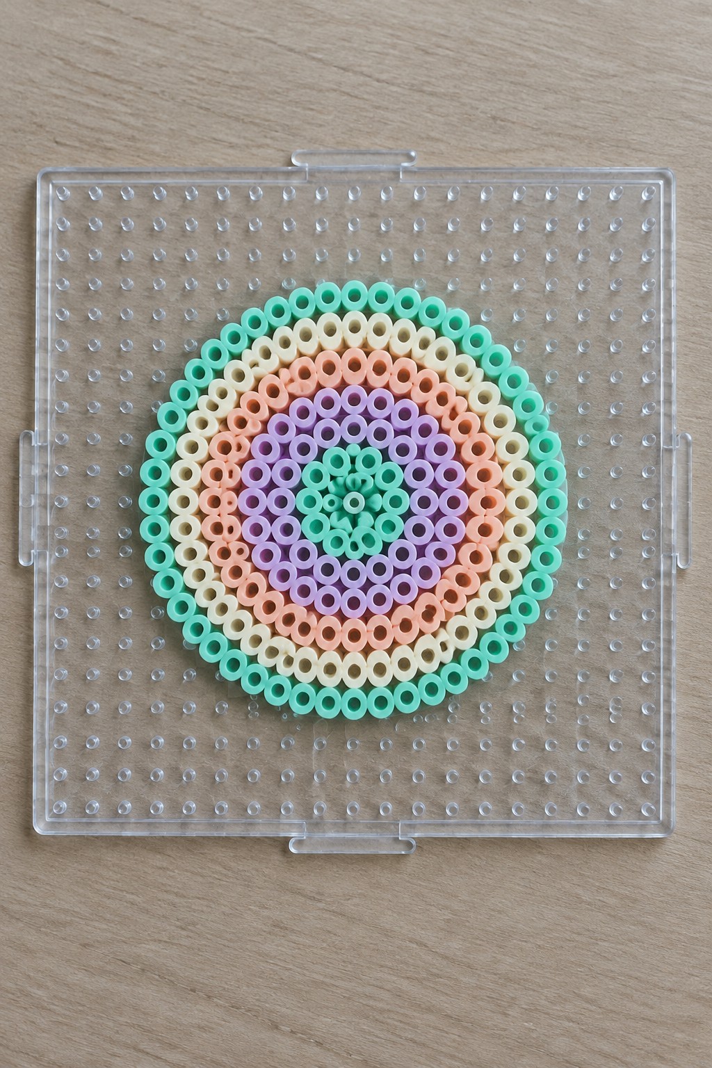 Pastel beads forming concentric circles on a transparent square pegboard.
