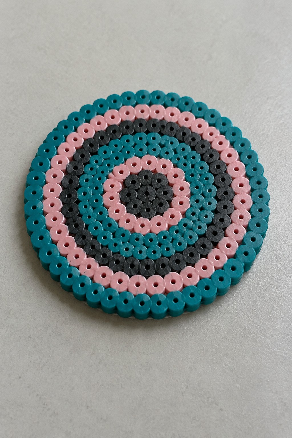 Circular perler bead coaster with concentric teal, pink, black, and gray rings