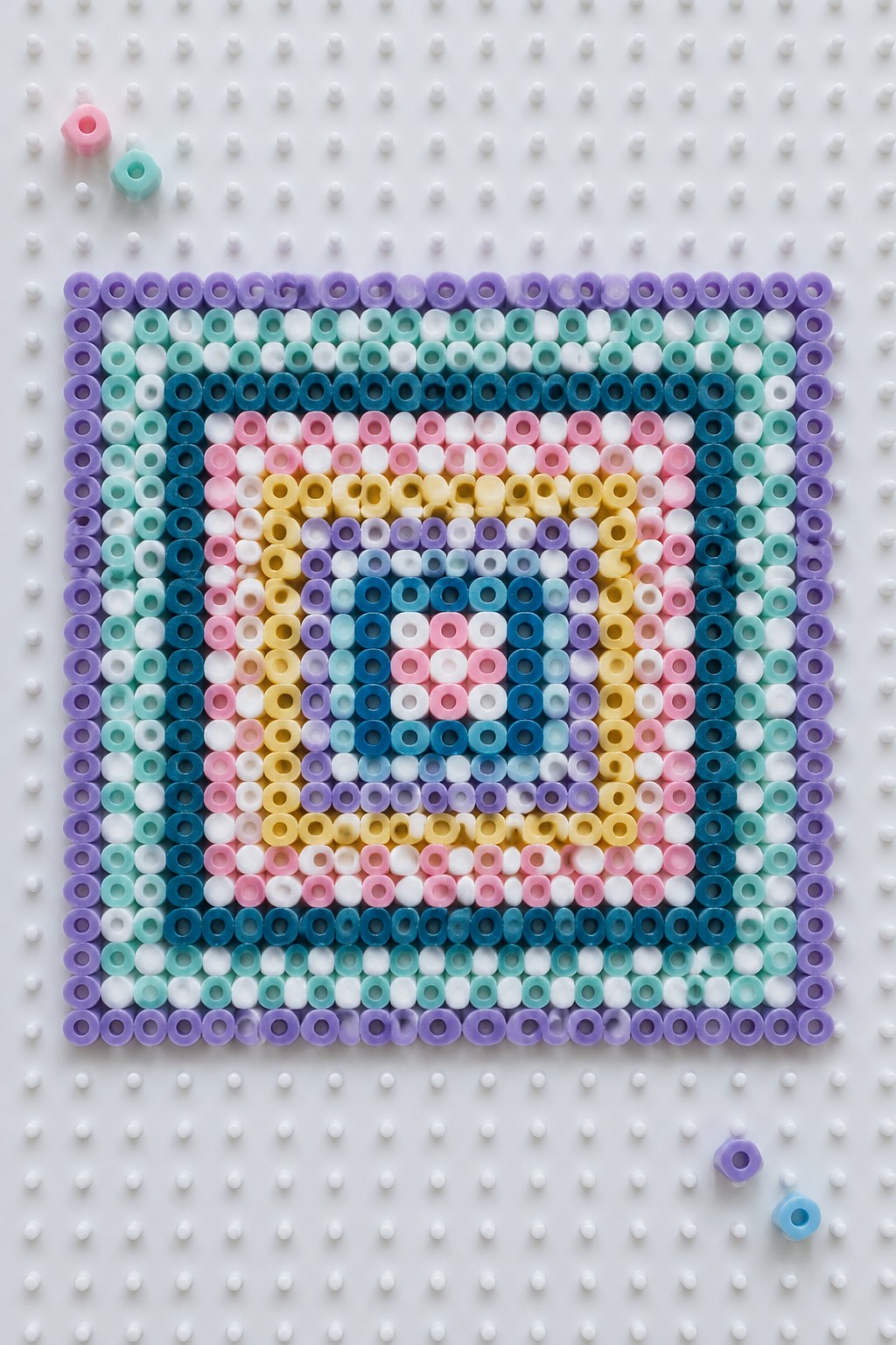 Colorful concentric square perler bead pattern on white pegboard with loose beads