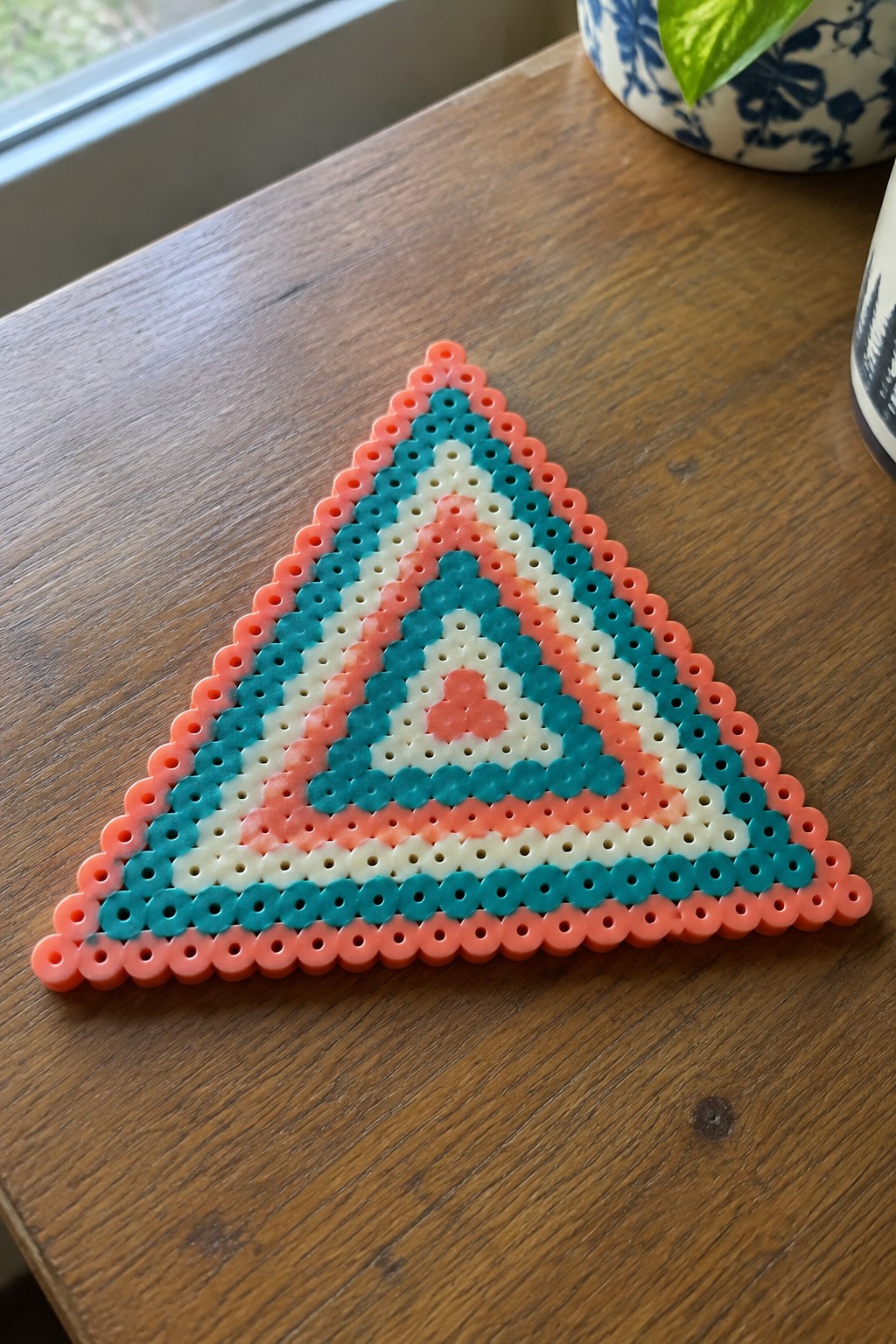 Triangular Perler bead art with layered coral, teal, and white concentric patterns on wood.