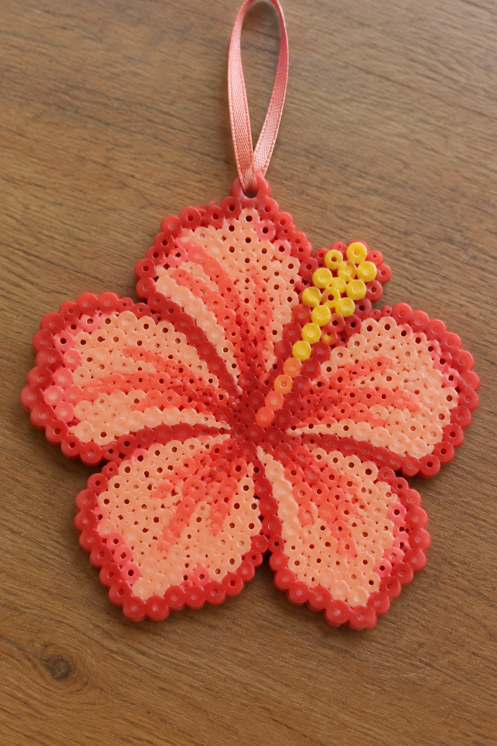 Pink and red perler bead hibiscus flower with yellow center and ribbon hanger on wood