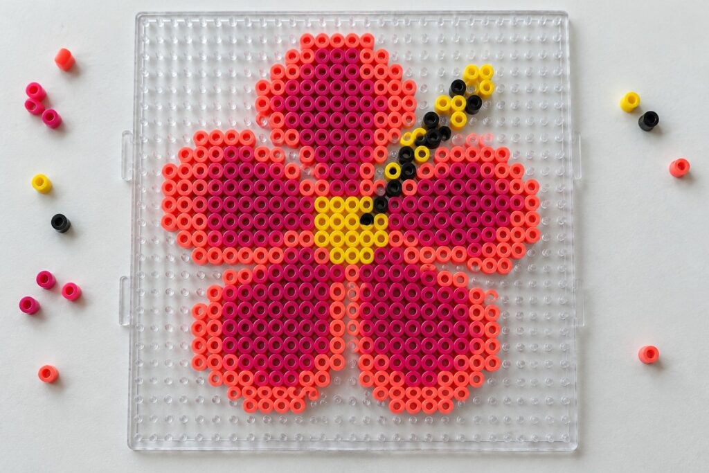 20 Tropical Hibiscus Perler Bead Patterns With Bold Petals Perler bead hibiscus flower pattern on a pegboard using pink, coral, magenta, yellow, and black beads.