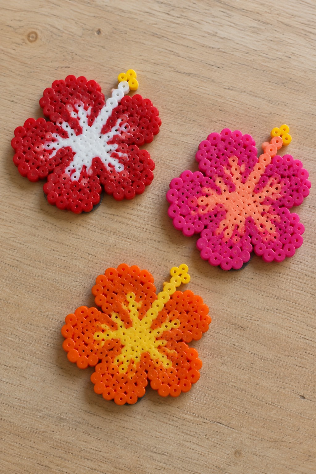 Three perler bead hibiscus flowers in red, pink, and orange on wood.