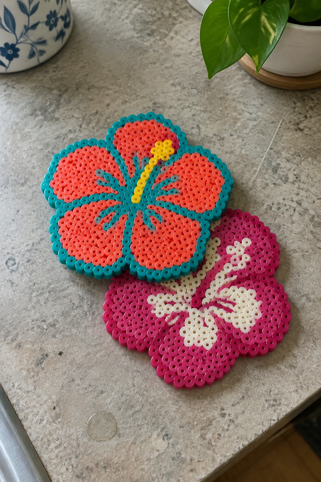 Two perler bead hibiscus coasters, orange and magenta, on a countertop.