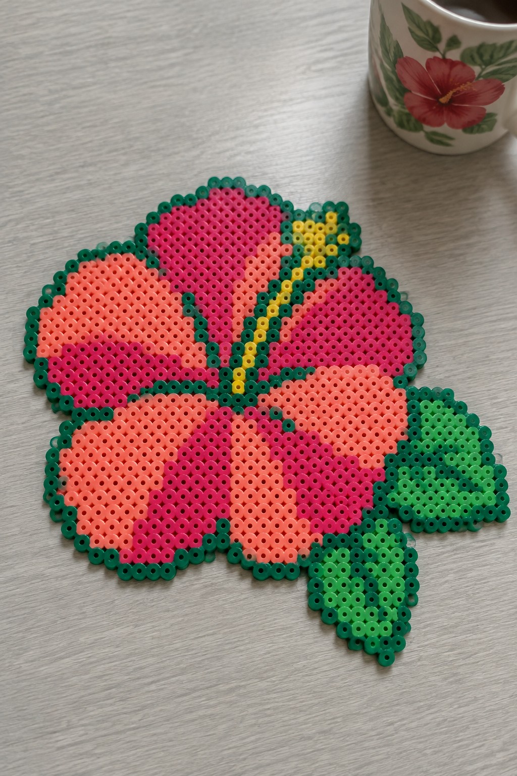 Perler bead hibiscus flower with pink and coral petals, yellow center, and green leaves on a pegboard.