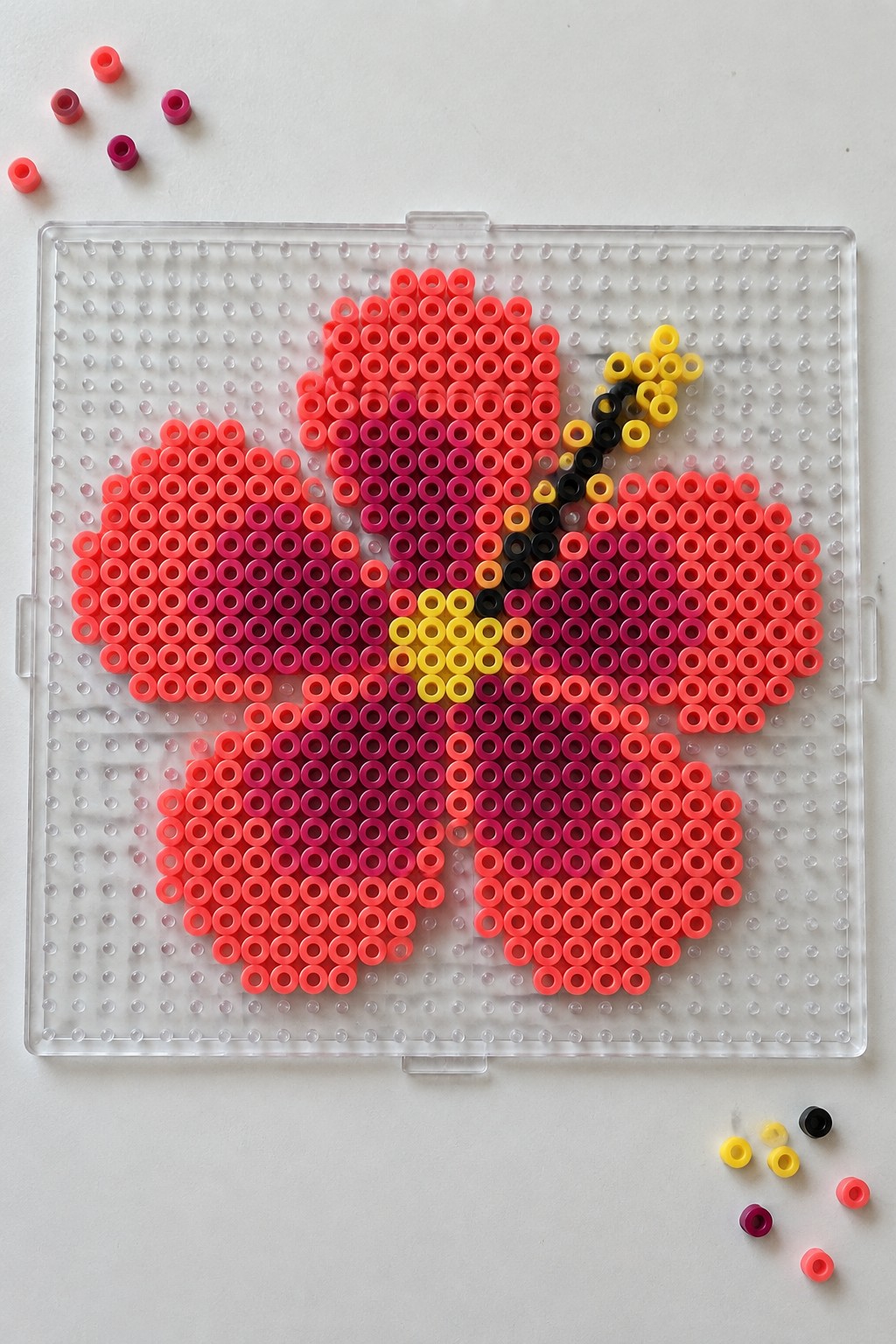 Perler bead hibiscus flower pattern on a pegboard using pink, coral, magenta, yellow, and black beads.