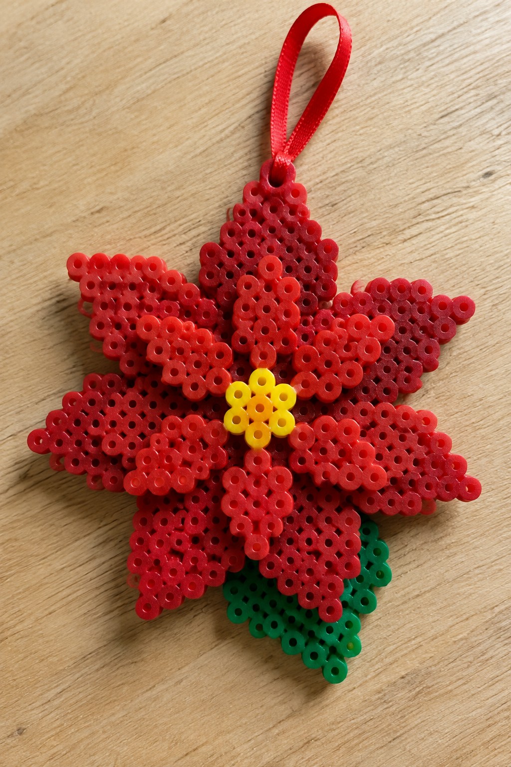 Red perler bead poinsettia ornament with yellow center, green leaves, red ribbon