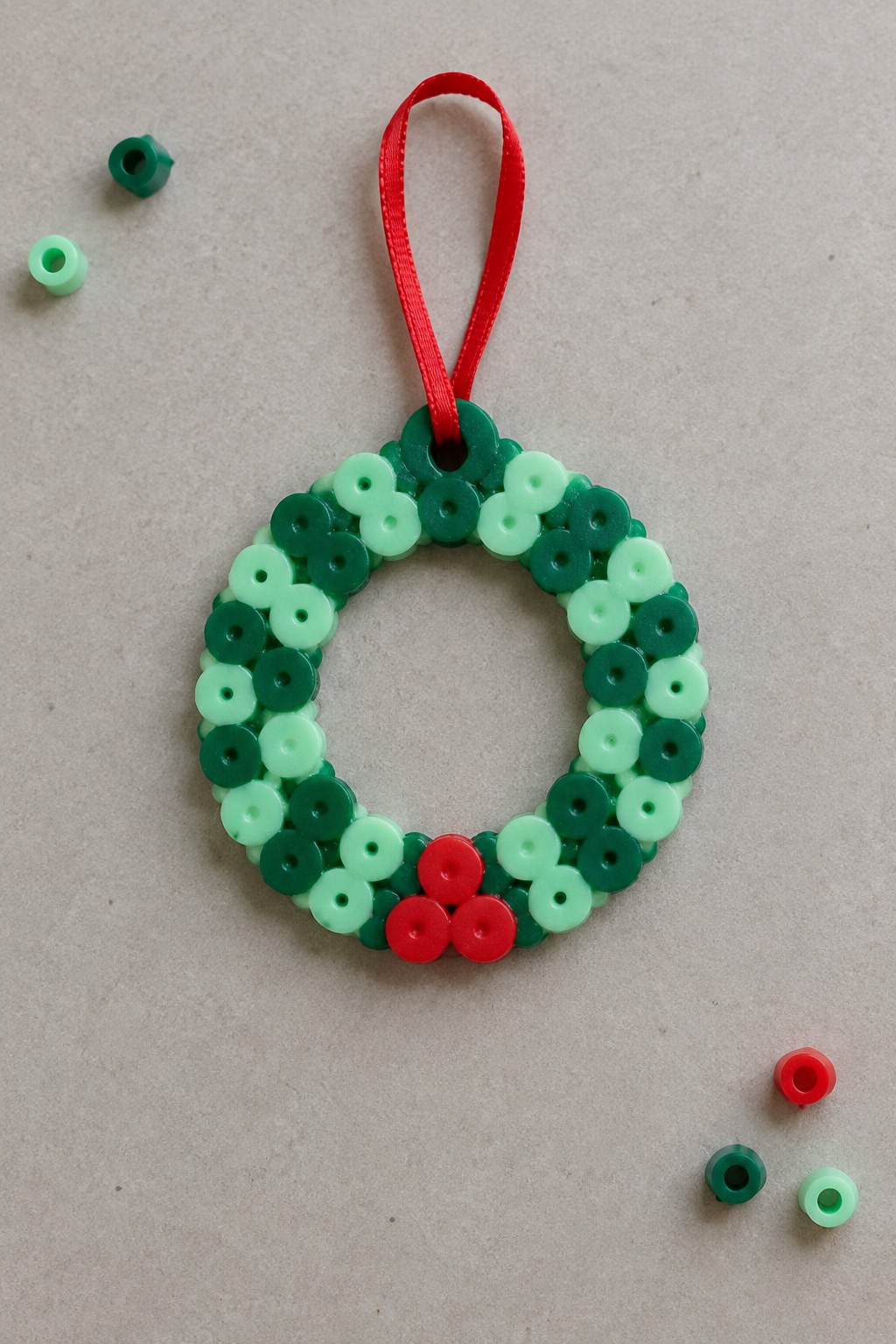 Perler bead Christmas wreath ornament made with multiple green shades, red berry beads at the bottom, and a red ribbon hanger.
