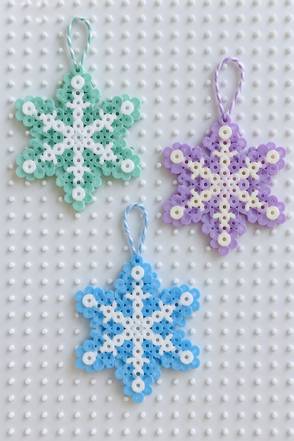 Three Perler bead snowflake ornaments on a pegboard in mint green, purple, and blue color schemes with white accents and hanging loops.
