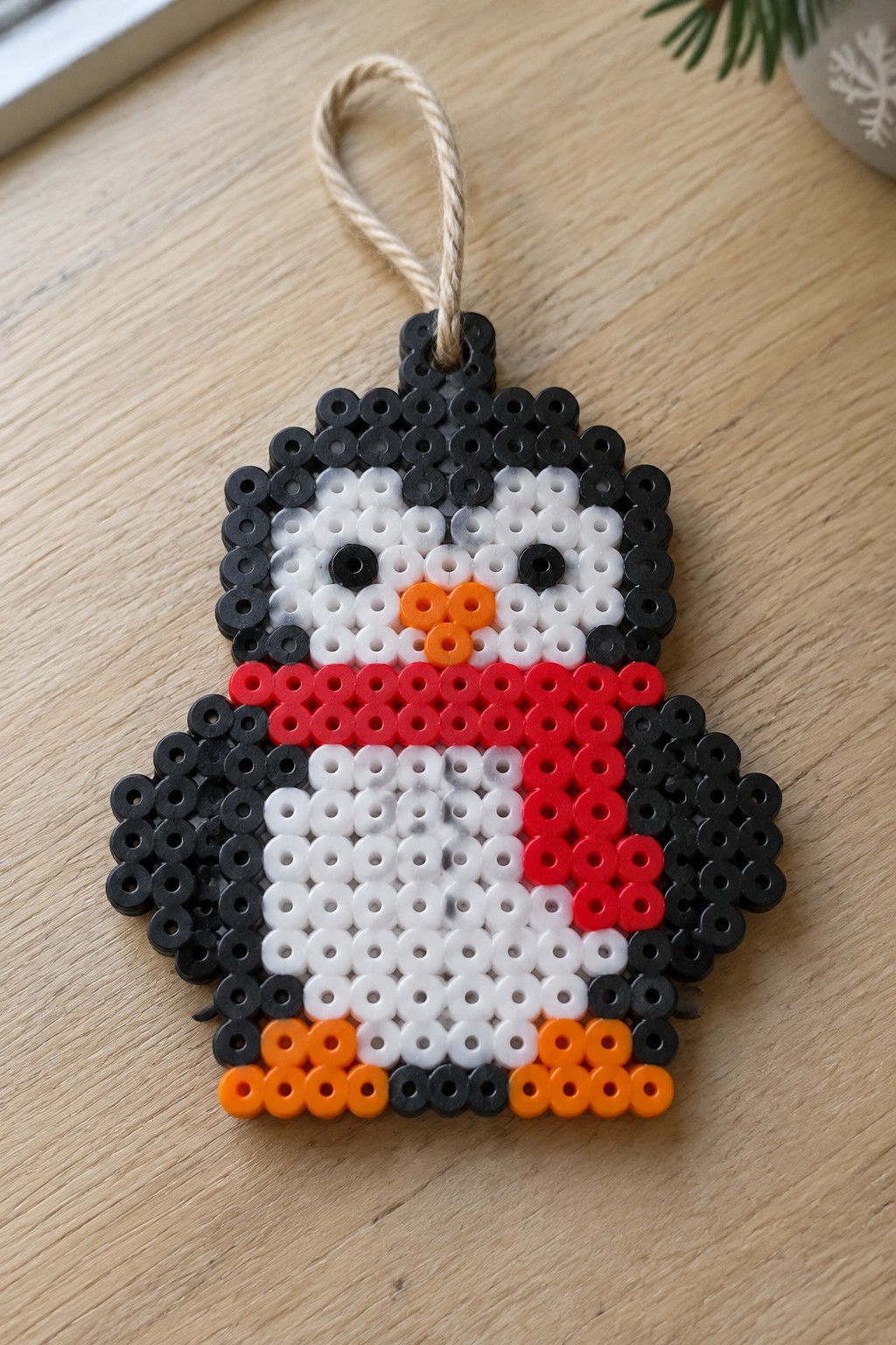 Perler bead penguin ornament with black body, white face, red collar, orange beak on wood.