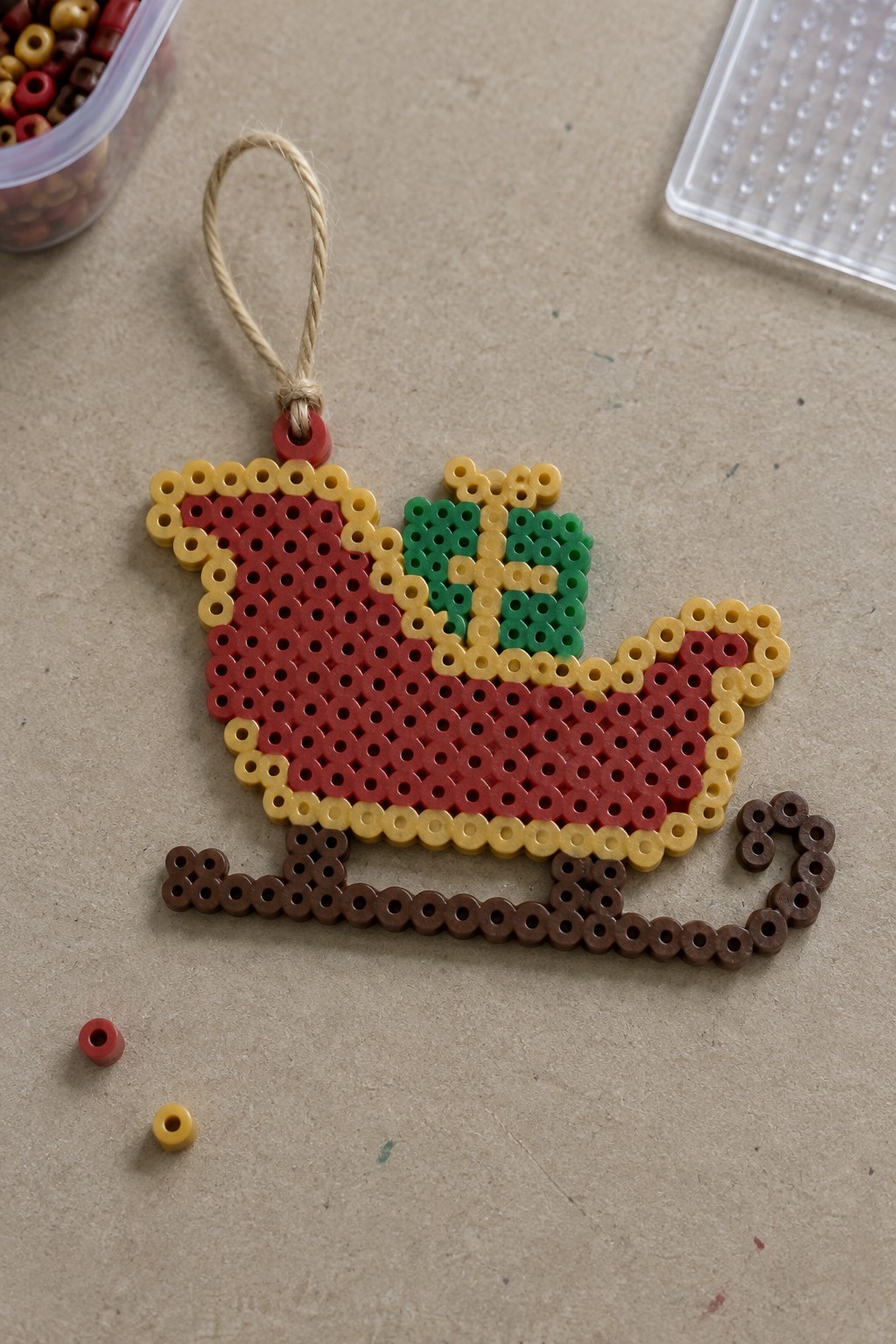 Perler bead Santa's sleigh ornament with red body, green present, and brown runners.