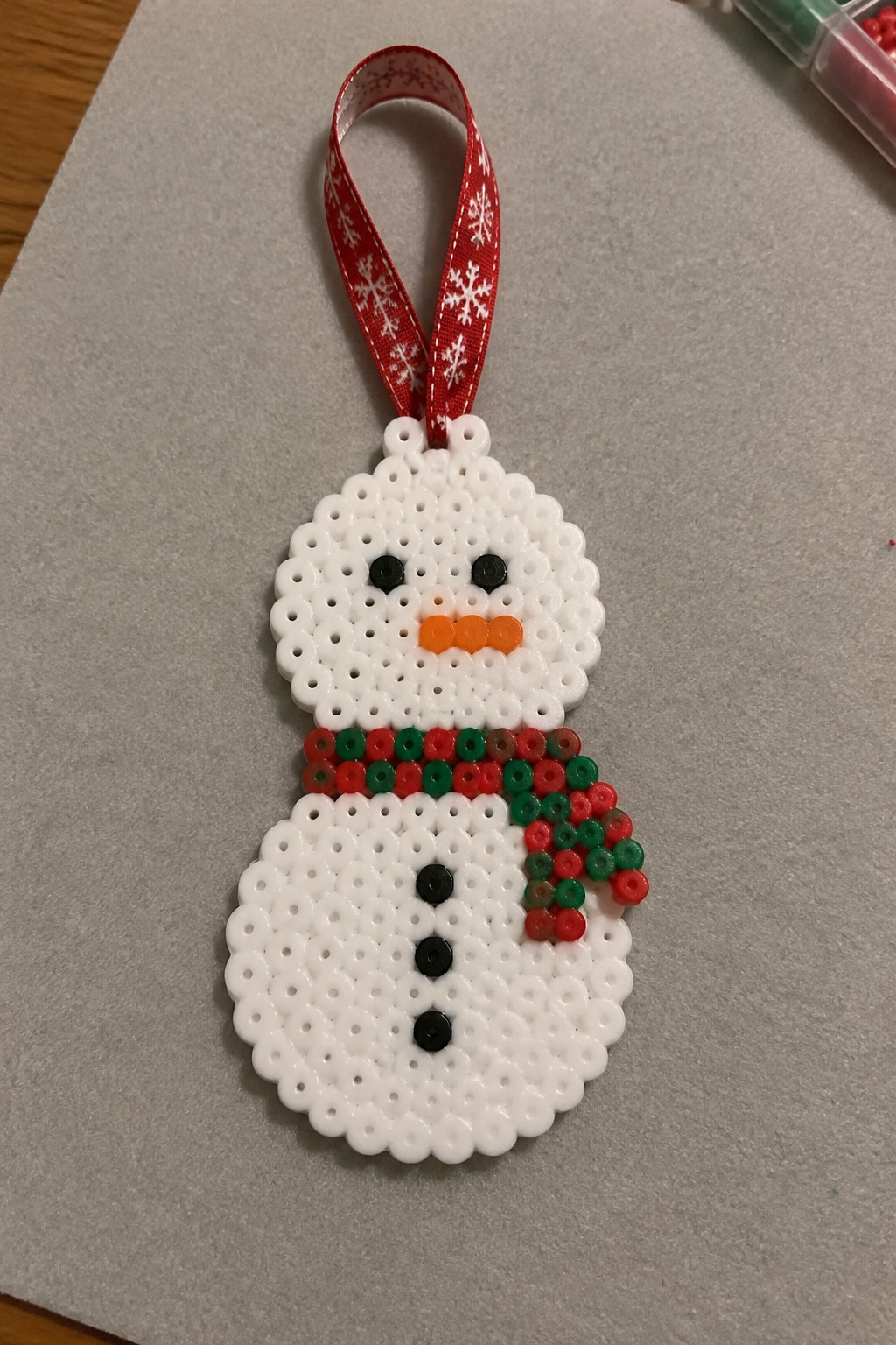A fused Perler bead snowman ornament with white body, black eyes and buttons, orange nose, red and green scarf, and red snowflake ribbon.