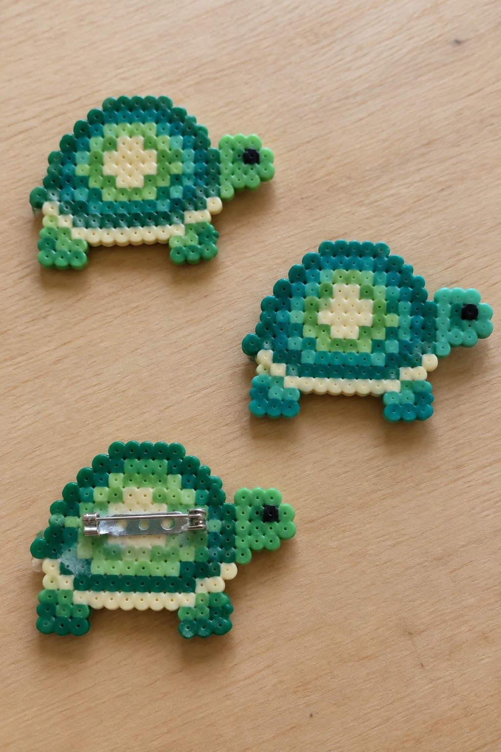 Three green perler bead turtles on wooden surface, one with metal pin