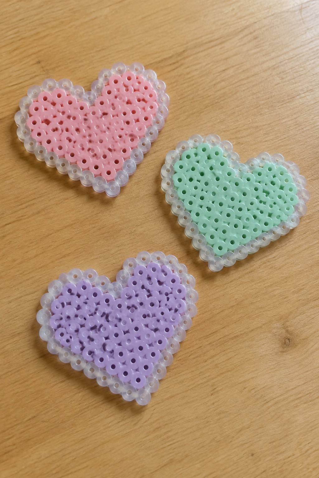 Three pink, green, and purple perler bead hearts with clear borders on wood