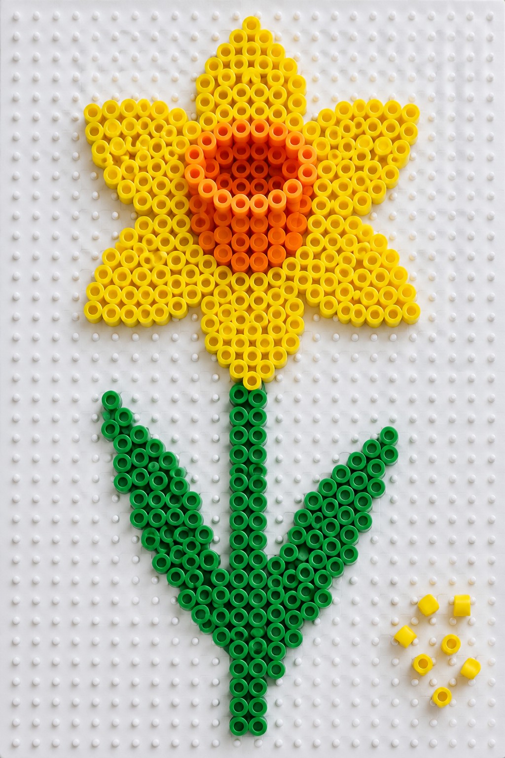 Perler bead flower with yellow-orange petals, green stem, and leaves on pegboard.
