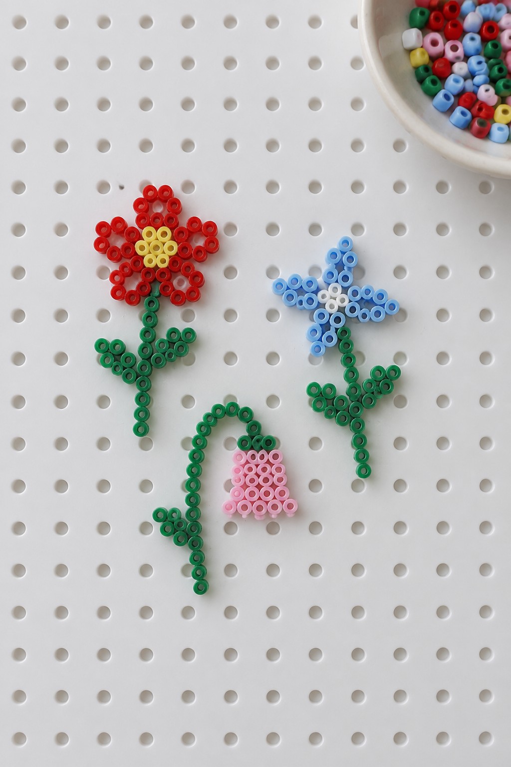 Three perler bead flowers with green stems on white pegboard beside bead bowl.