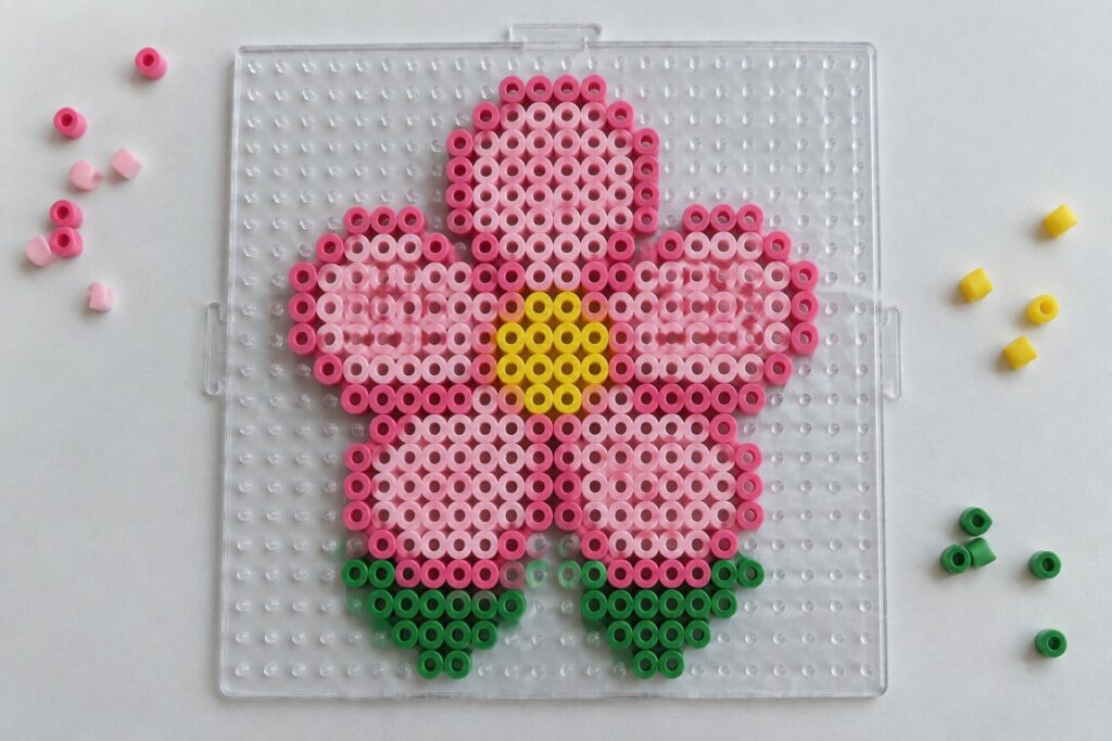 Pink flower Perler bead pattern with yellow center and green leaves on a clear pegboard.