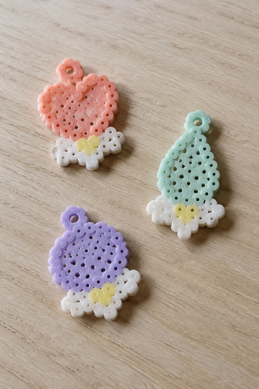 Three pastel perler bead ornaments—a pink heart, purple circle, and mint tree—on wood.