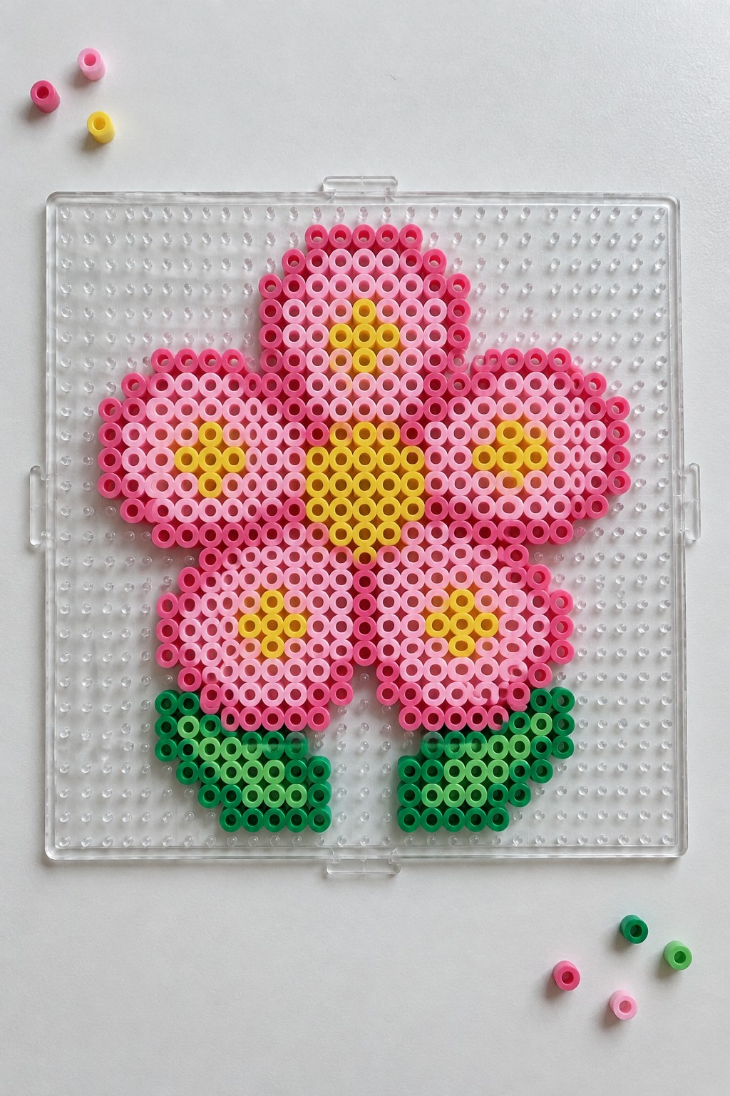 Pink flower Perler bead pattern with yellow center and green leaves on a clear pegboard.
