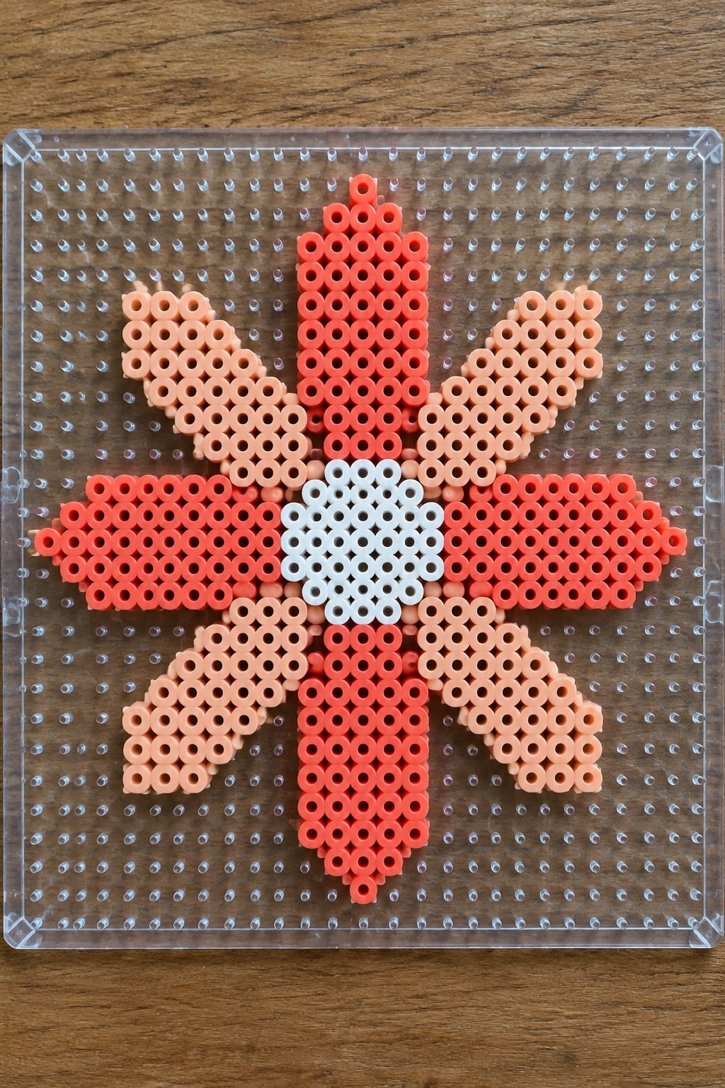 Symmetrical perler bead flower in red, orange, peach and white on clear pegboard