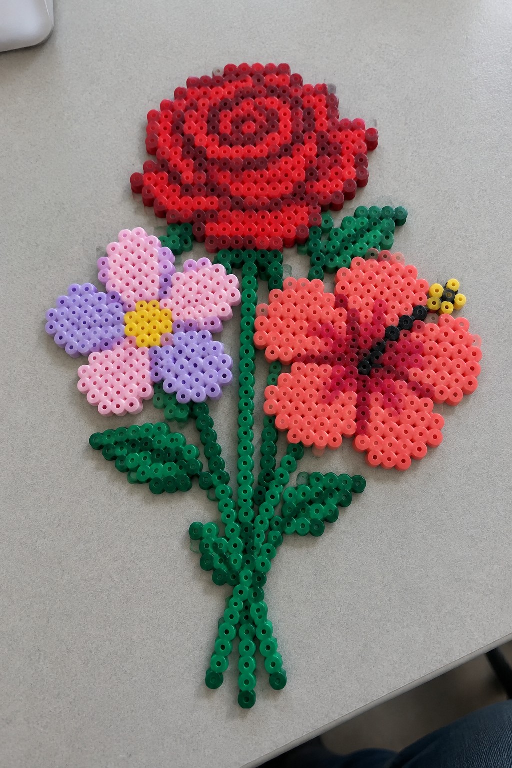 Perler beads forming red rose, purple flower, orange bloom with bee, green stems