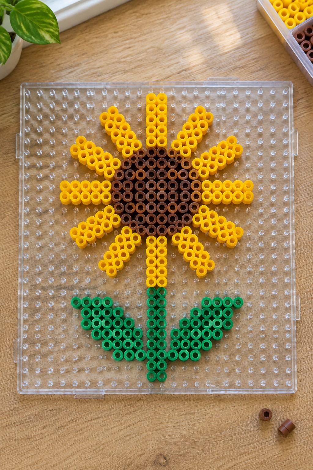 Perler bead sunflower with yellow petals, shaded brown center, green stem and leaves on a clear pegboard.