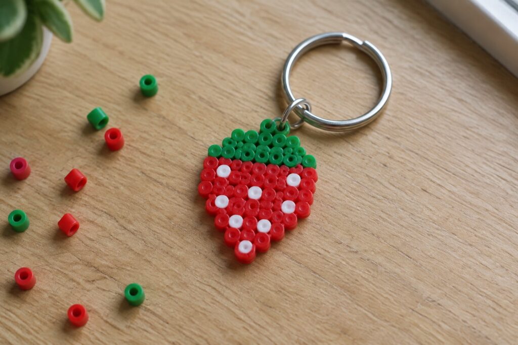 21 Mini Perler Bead Designs for Tiny Keychains and Charms Strawberry Perler bead keychain with red body, white seeds, green top on wood.